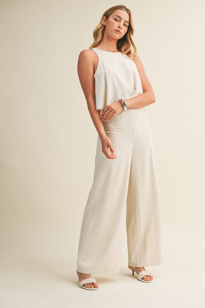 Woman wearing a cream linen sleeveless jumpsuit with wide legs and white sandals.