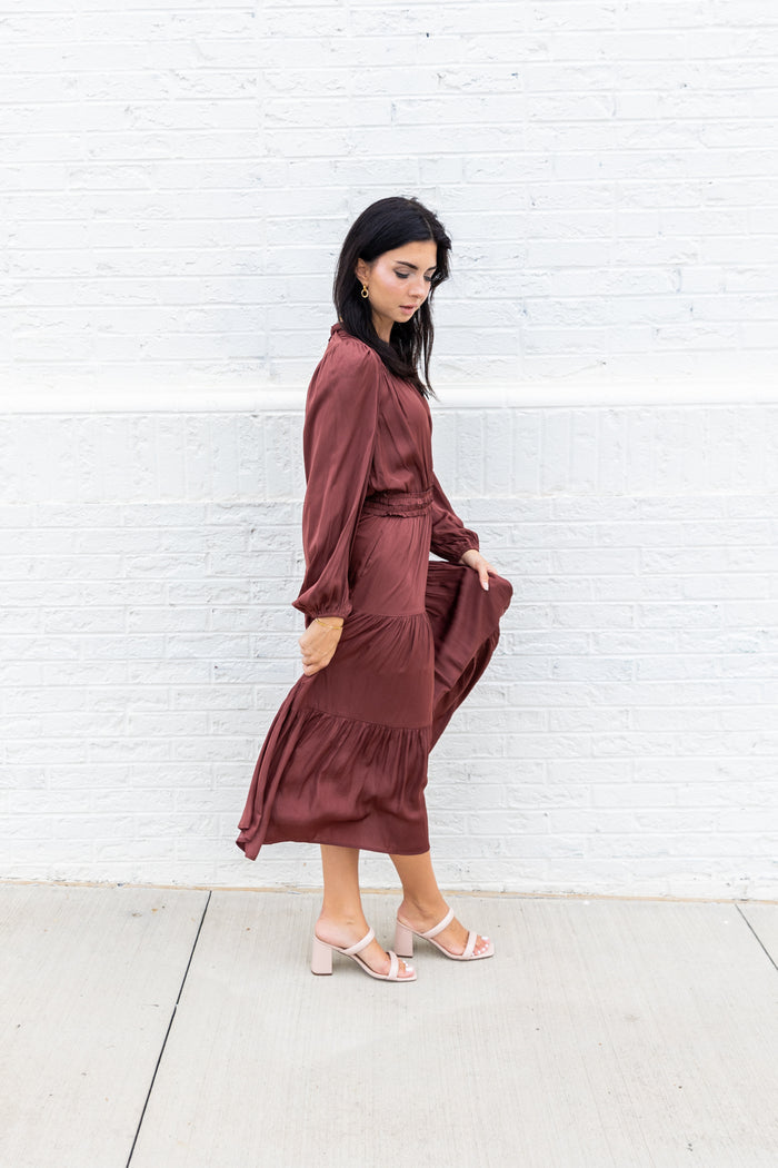 Burgundy maxi dress with long sleeves, cinched waist, and tiered skirt on model against white brick wall.