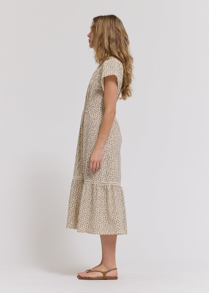 Beige midi dress with small floral print, short sleeves, and tiered skirt hem on model with long wavy hair.