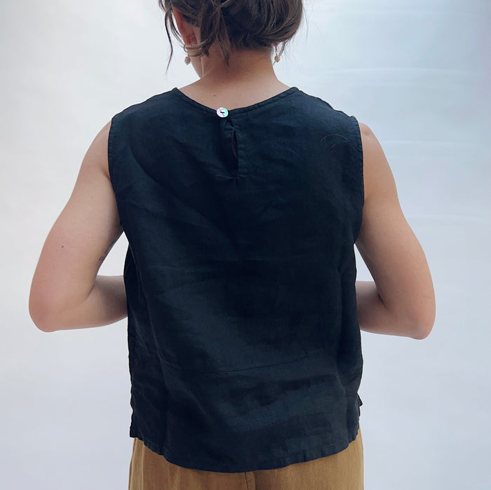 Back view of model wearing Bryn Walker black basic linen tank, showing single back button closure at neckline, sleeveless cut, relaxed boxy silhouette, side slits, and black color, styled with camel wide-leg pants