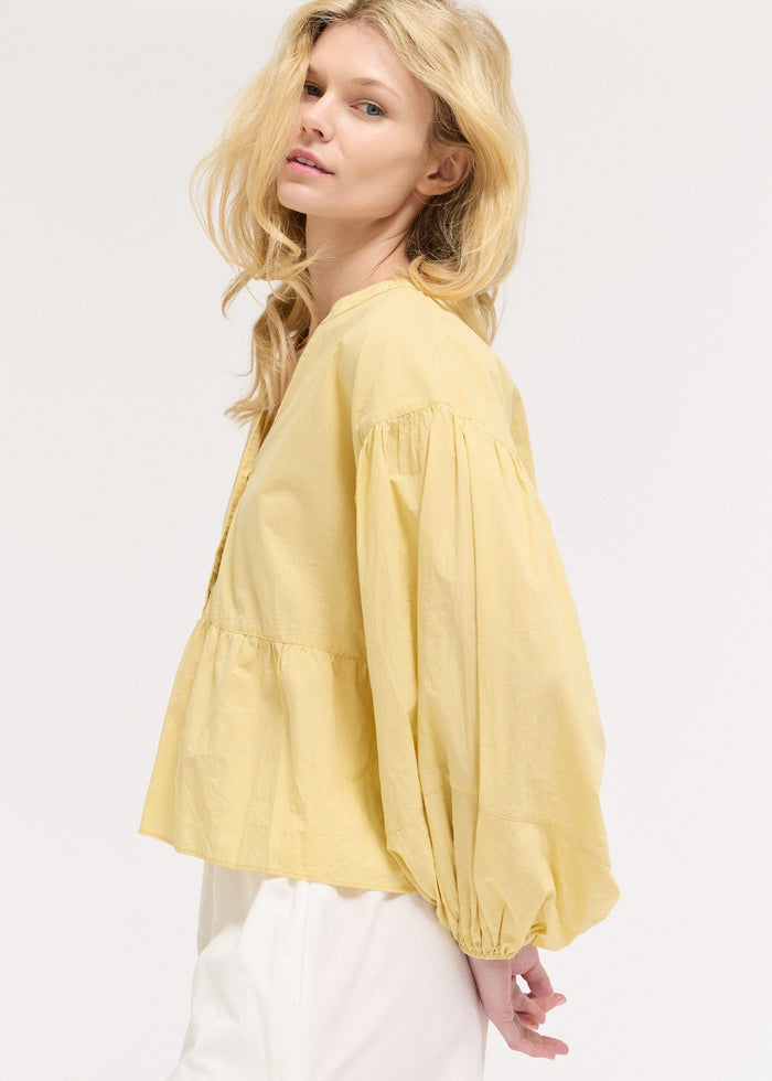Woman wearing pale yellow blouse with voluminous balloon sleeves and white pants, shown from side profile.