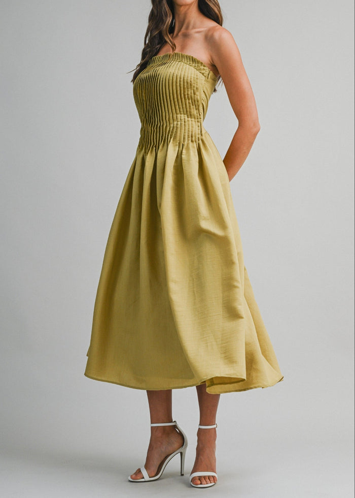 Olive-yellow strapless midi dress with pleated bodice and full skirt, worn with white heeled sandals.