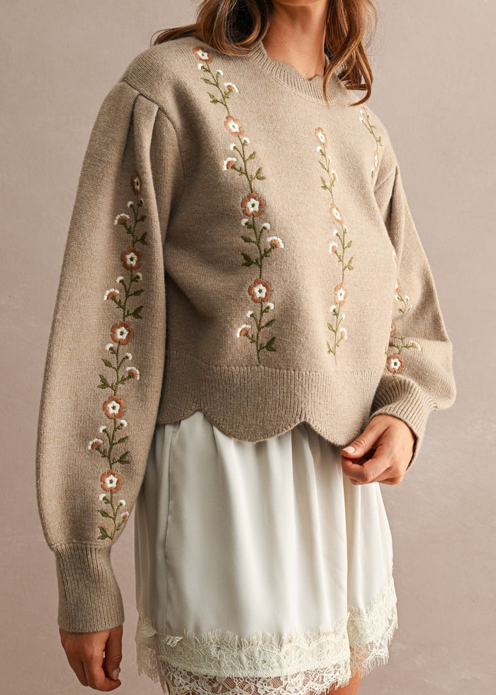 A woman wearing a beige knit sweater with floral embroidery on the front and sleeves, paired with a white skirt, standing against a neutral beige background.