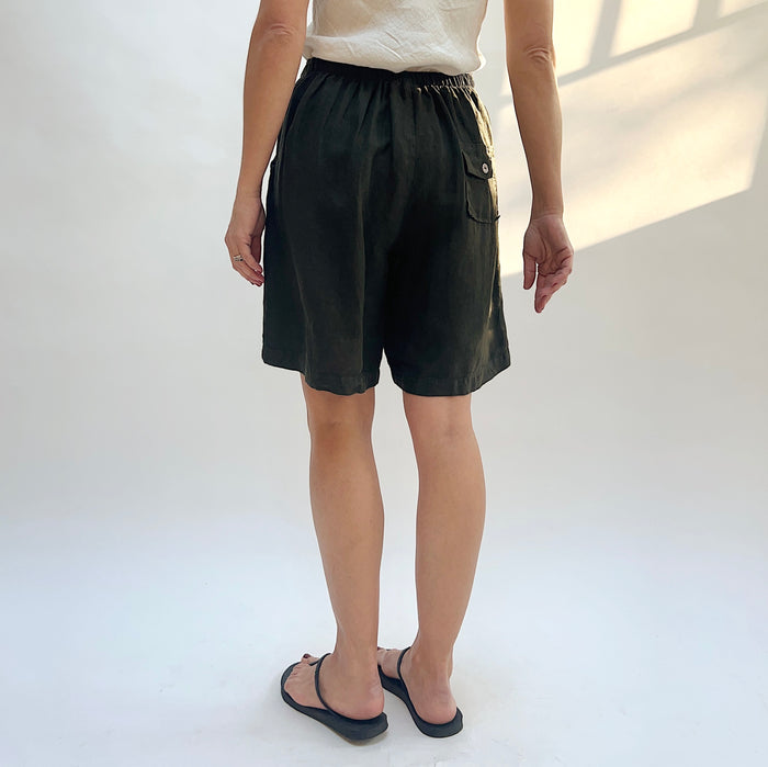Back view of model wearing Bryn Walker peppercorn linen sasha short, showing elastic waistband, back patch pocket with button, relaxed wide-leg bermuda length, and deep charcoal color, with black thong sandals