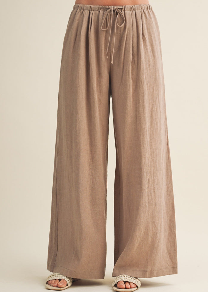 Beige wide-leg pants with drawstring waist and relaxed fit, shown on model wearing white slides.