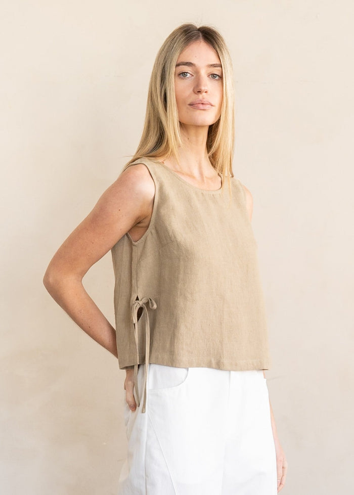 Woman wearing a beige sleeveless linen top with side tie detail and white pants.