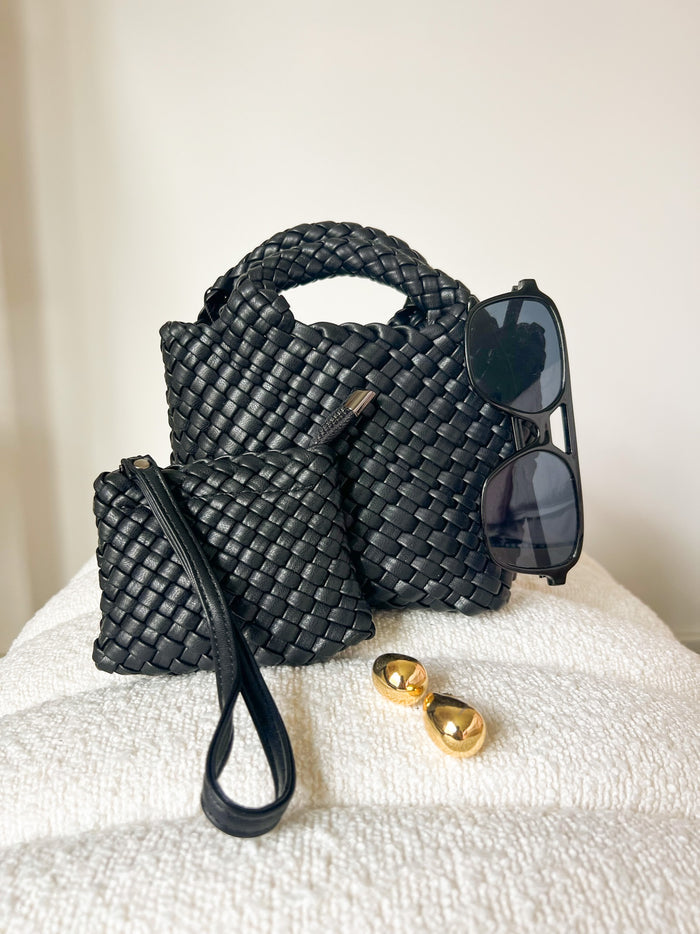 Black woven handbag with dual handles, matching woven pouch, sunglasses, and gold earrings displayed on cream fabric.