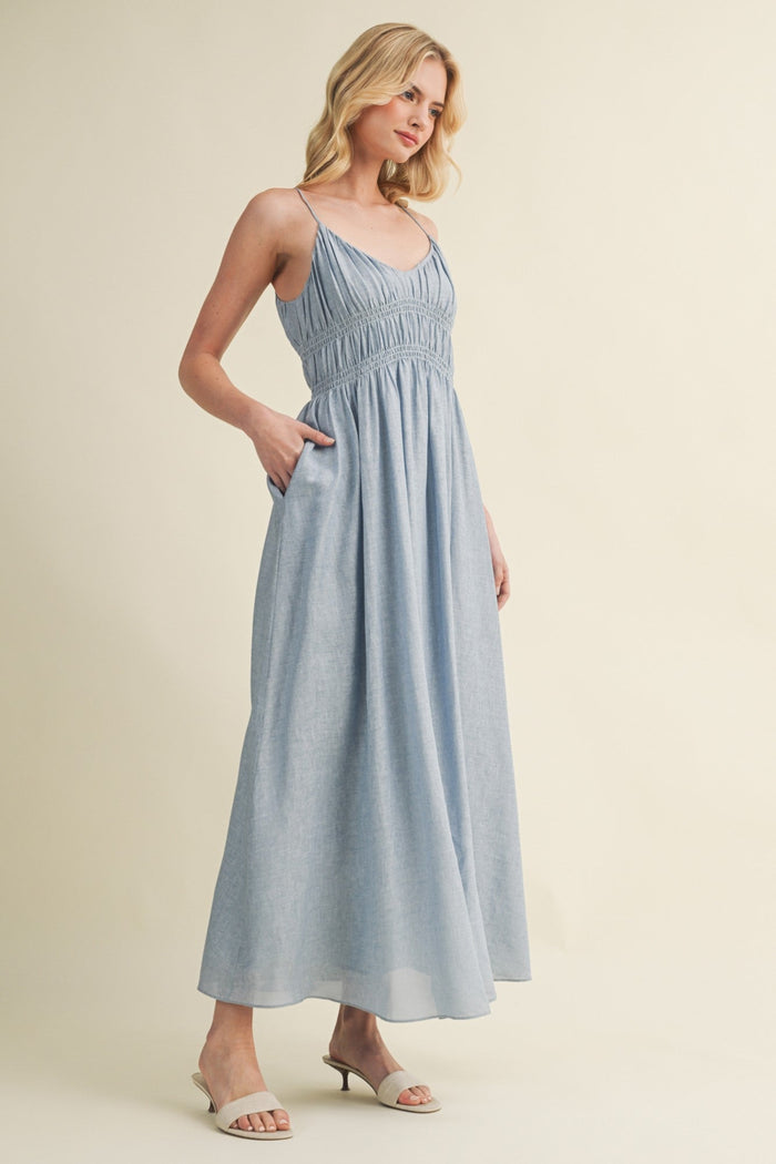 Blue-gray sleeveless maxi dress with ruched empire waist and side pockets on model wearing neutral sandals.
