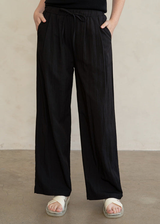 A person wearing loose black wide-leg pants with an elastic drawstring waist, paired with a black sleeveless top and white slide sandals, standing against a plain light beige wall.
