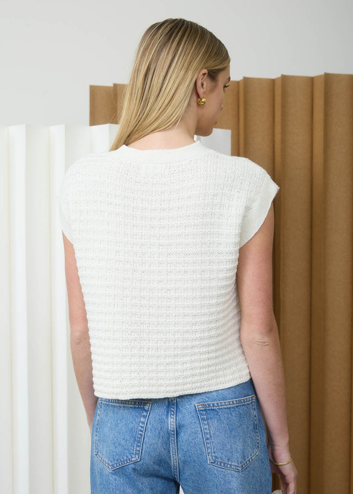 Person wearing a white sleeveless top and blue jeans standing in front of a white wall with a brown curtain.