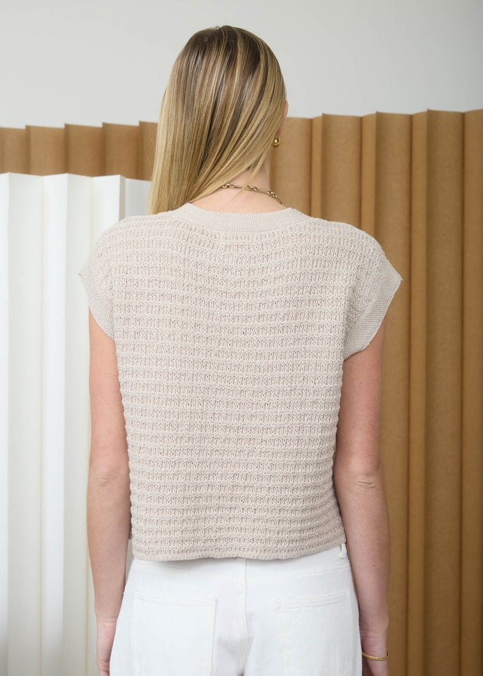Woman wearing a beige knitted top and white pants against a neutral background