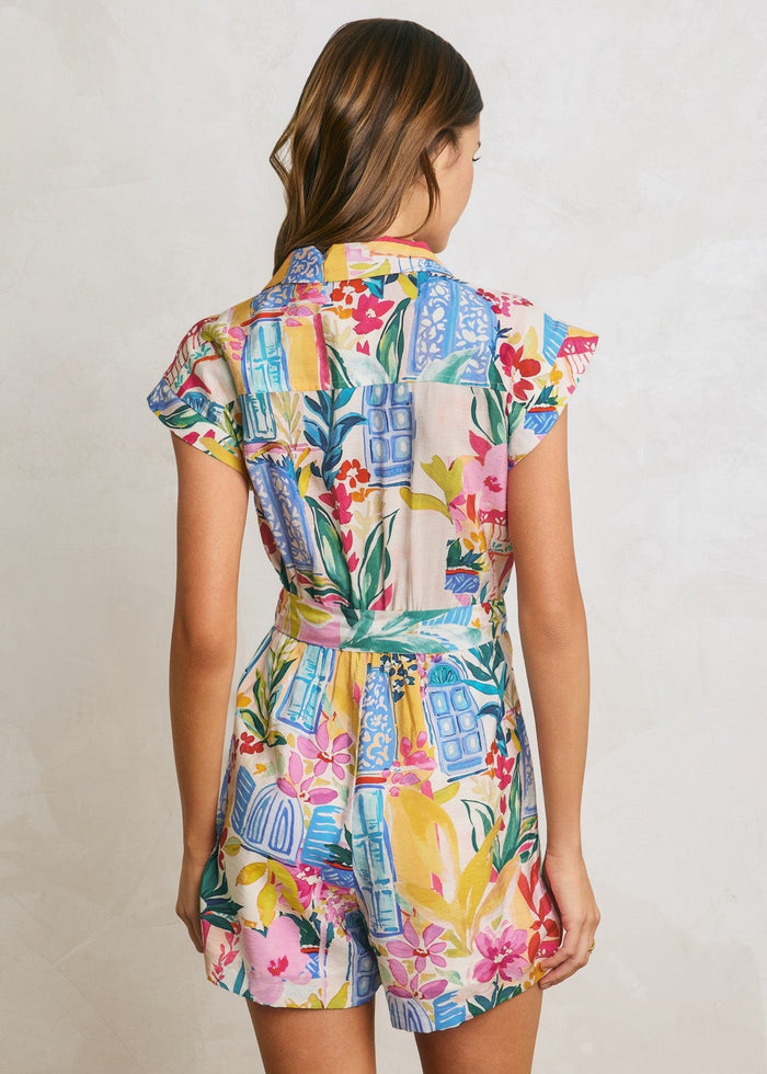 Woman wearing a colorful floral and geometric patterned button-down romper with short flutter sleeves and a belted waist.