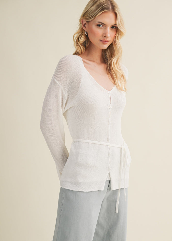 Woman wearing a cream knit cardigan with button front and tie waist over gray pants.
