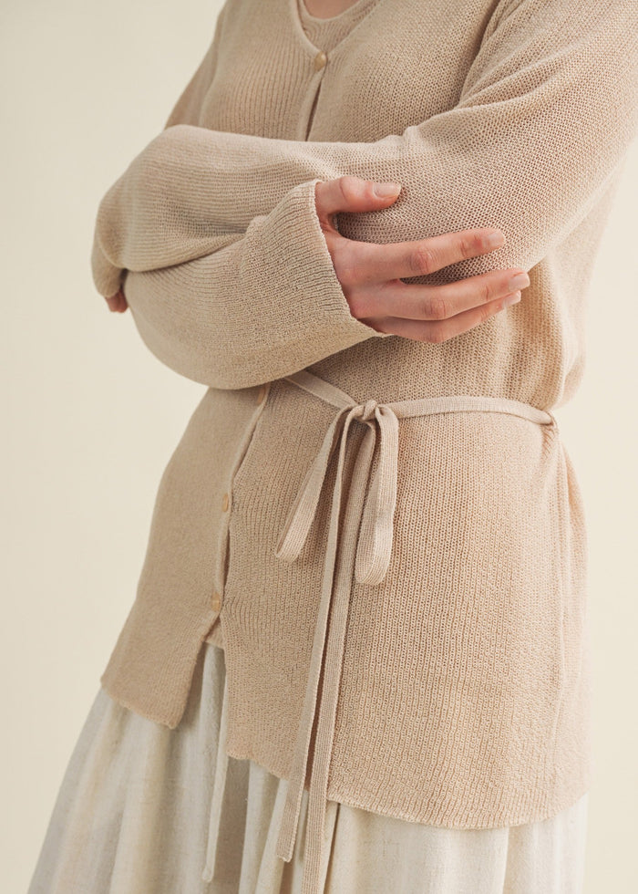 Beige knit cardigan with button front and tie waist, worn open over a white skirt.