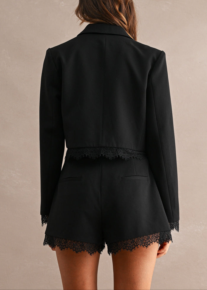 A woman shown from the back wearing a black cropped blazer and matching black shorts with lace trim, against a plain warm beige studio background.