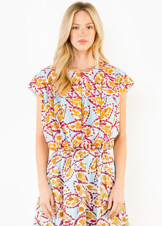 A woman modeling a short-sleeve wrap dress with a colorful abstract print on a light blue background. The dress features yellow, orange, and magenta patterns, and is photographed against a plain white studio background.