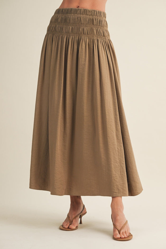 Tan a-line maxi skirt with smocked waistband and ankle-length hem on model wearing strappy sandals.