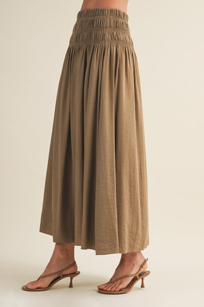 Tan a-line maxi skirt with smocked waistband and pleated detail, paired with brown kitten heels.
