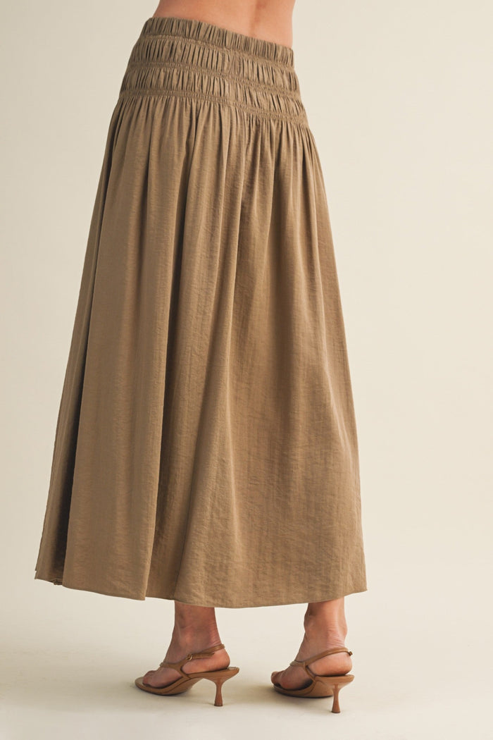 Tan a-line maxi skirt with shirred elastic waistband, shown from back with brown heeled sandals.