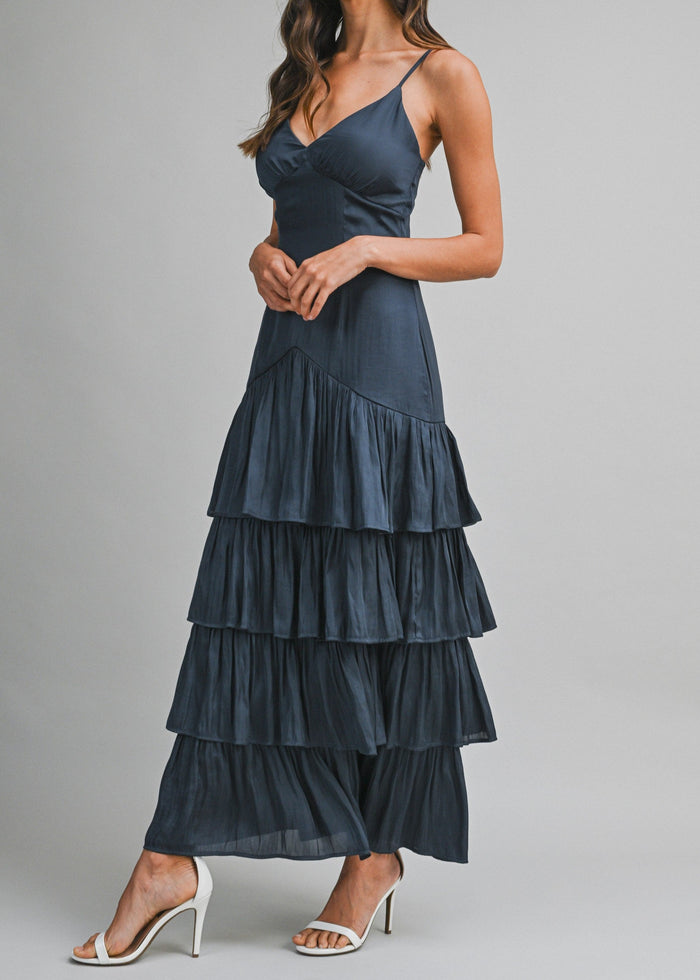 A woman modeling a dark slate blue tiered maxi dress with spaghetti straps and white high-heeled sandals against a solid gray studio background.
