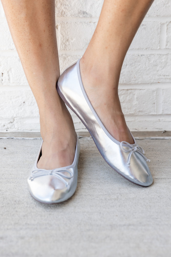 Silver metallic slip-on ballet flats with bow detail worn on feet against white brick background.