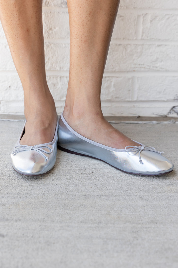 Silver metallic ballet-style flat shoes with front bow detail worn on feet.