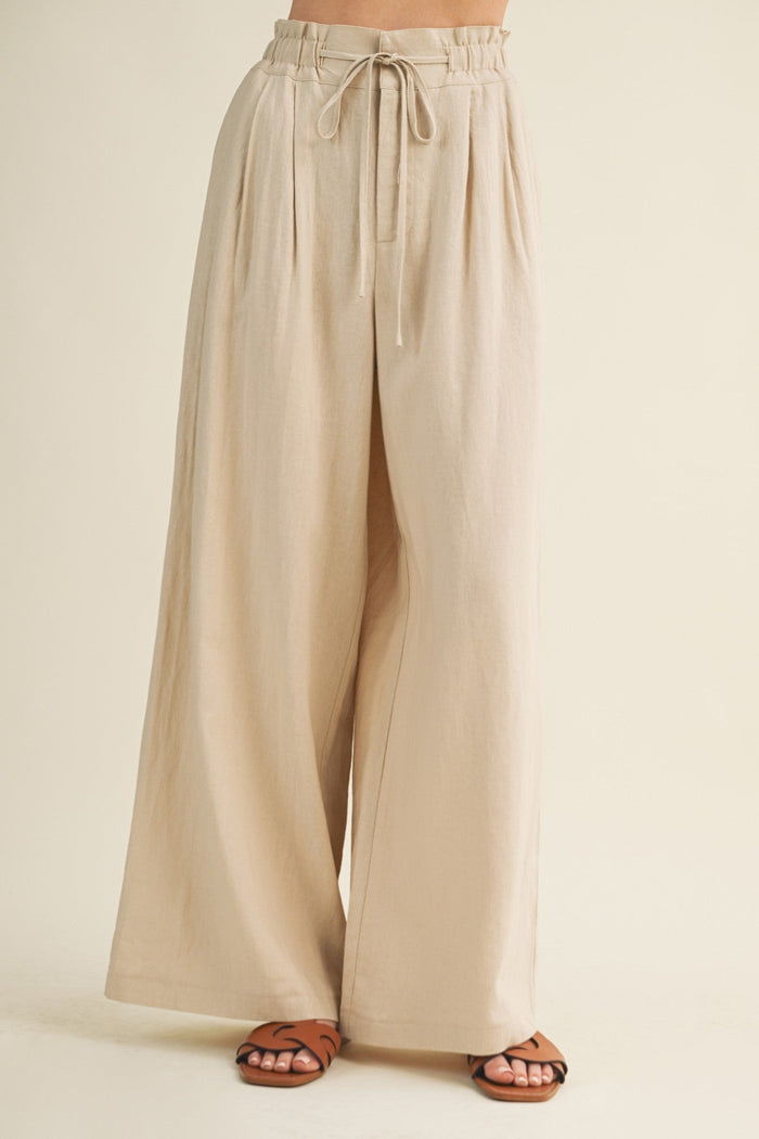 Beige wide-leg pants with elastic waistband and drawstring tie, shown on model wearing brown sandals.