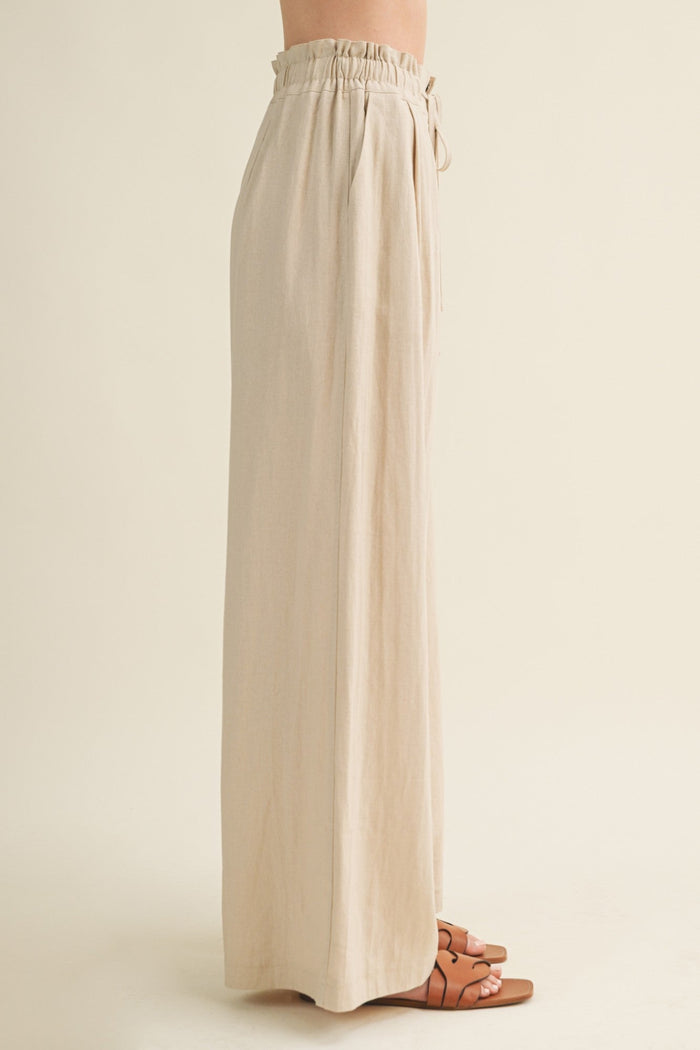 Beige wide-leg pants with elasticized waist and tie closure, shown from side view on model wearing brown sandals.
