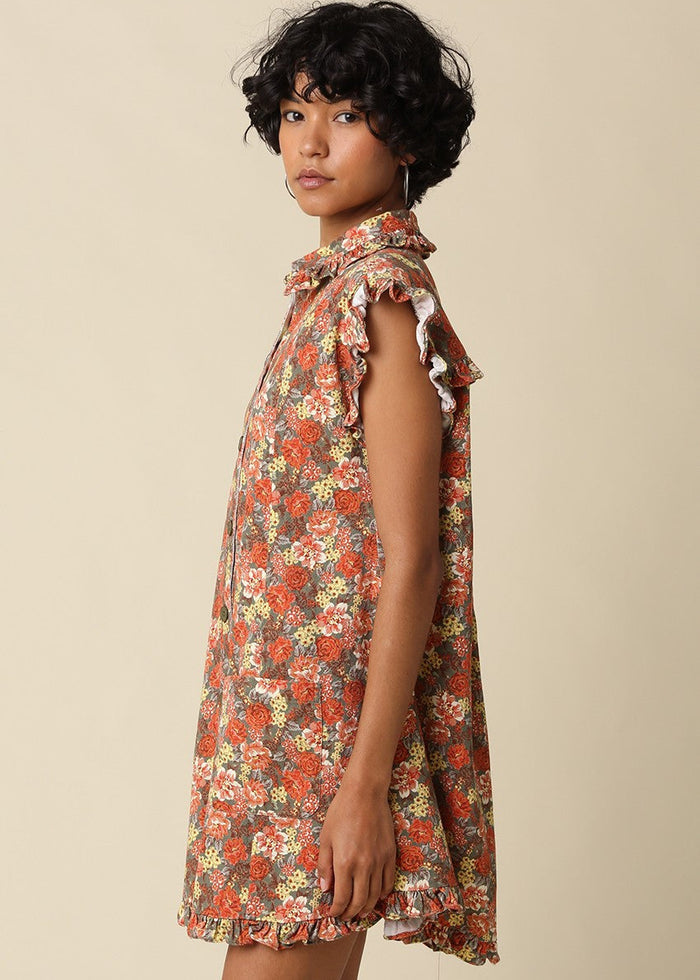A woman wearing a sleeveless, high-neck mini dress with a dense floral print in red, orange, and yellow on a beige background, photographed against a solid light beige studio backdrop.