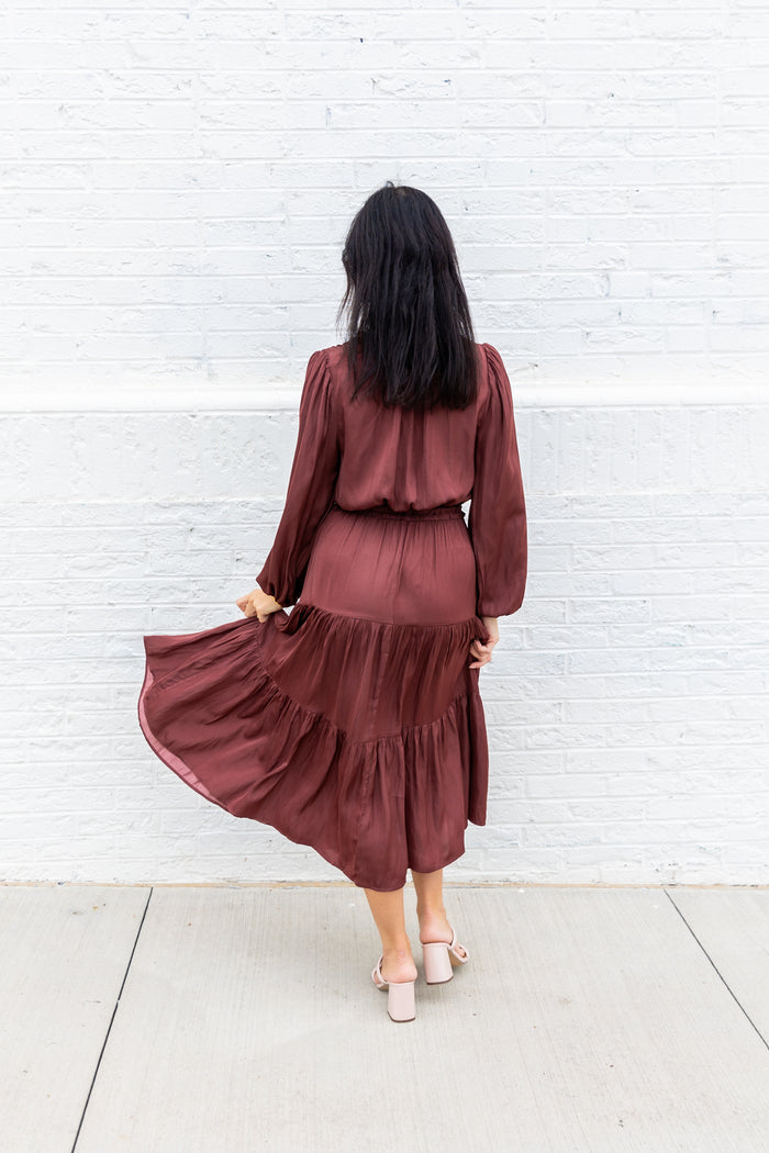 Woman wearing burgundy maxi dress with long sleeves and tiered skirt, shown from behind against white brick wall.