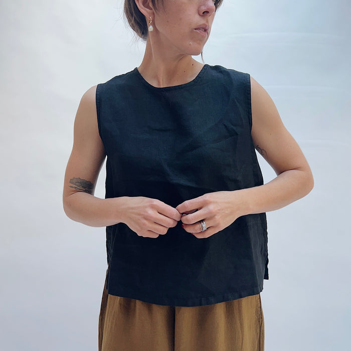 Full front view of model wearing Bryn Walker black basic linen tank with hands at hem, showing crew neckline, sleeveless cut, relaxed boxy fit, side slits, and black color, styled with camel wide-leg pants