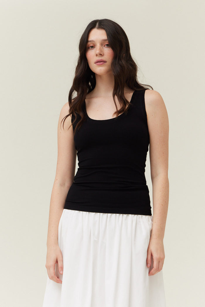 Black ribbed tank top with round neckline and sleeveless cut worn over white skirt.