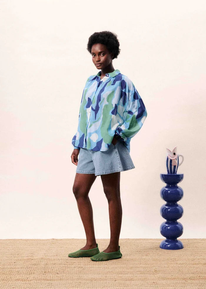 Person wearing a colorful shirt and shorts standing next to a blue vase on a wooden floor.