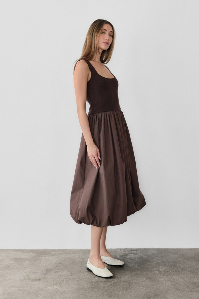 Brown sleeveless midi dress with fitted bodice and full gathered skirt, shown on model with white slip-on shoes.