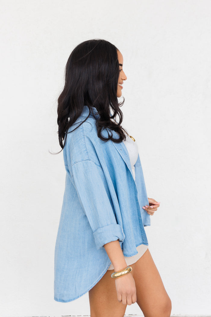 Light blue chambray button-up shirt with rolled sleeves worn over white undergarment, shown on model in profile view.