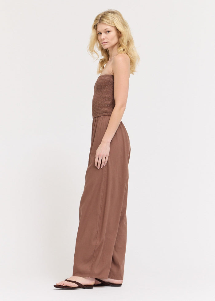 Woman wearing a taupe strapless jumpsuit with a smocked bodice and wide-leg pants with cocoon hem.