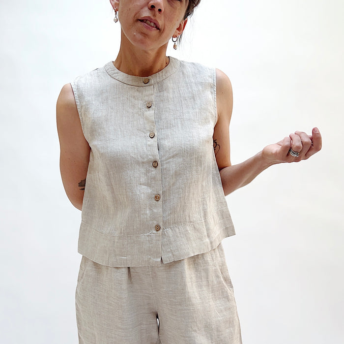 A close up shot of a person holding one arm behind her back wearing a matching button front linen tank in beige with beige linen pants.