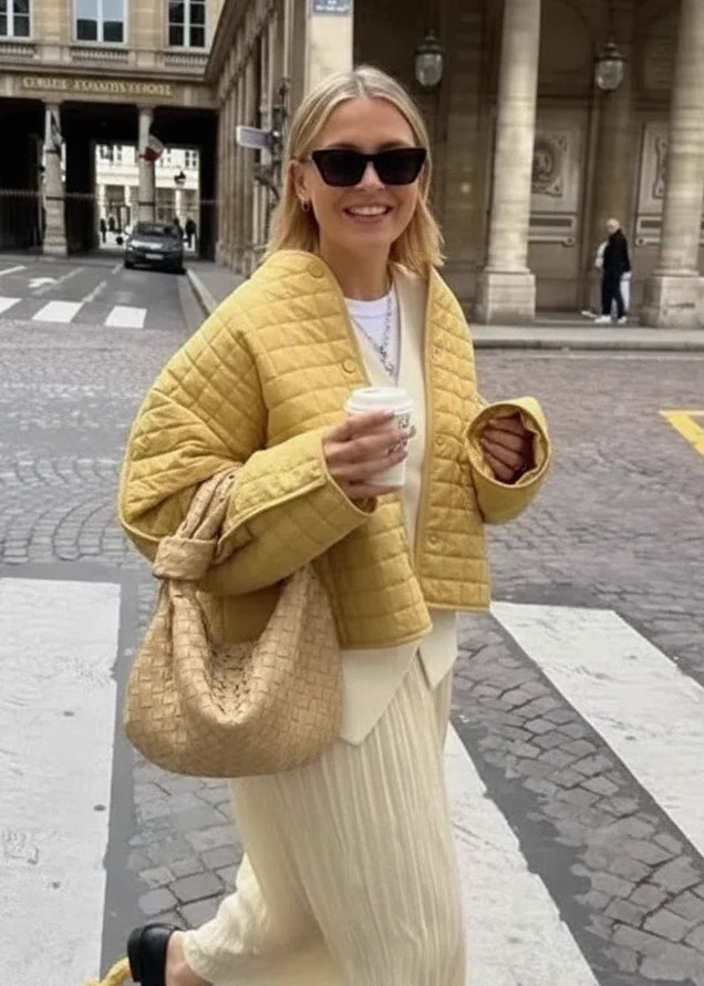 Woman wearing a yellow quilted jacket over a white top, styled with sunglasses and a matching quilted handbag.