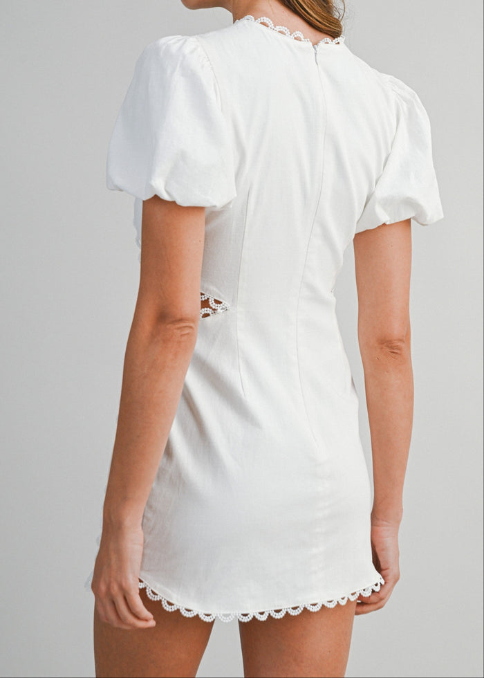 White mini dress with short puffed sleeves, lace trim neckline and hem, and distressed cutout details on back.