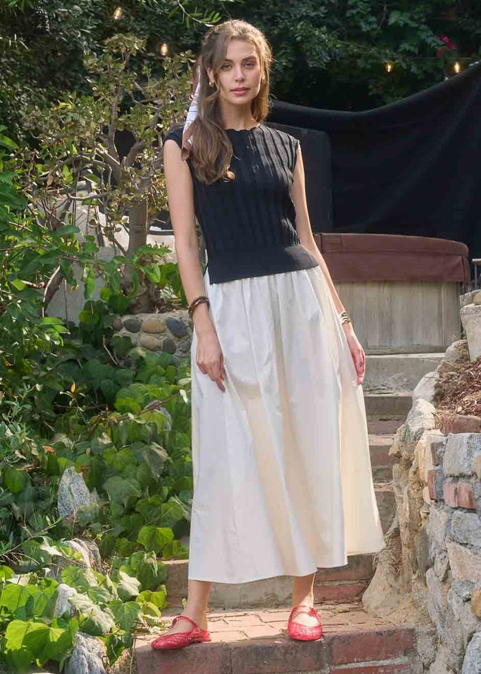 Woman wearing black sleeveless top with white midi skirt and red sandals, standing in a garden setting.