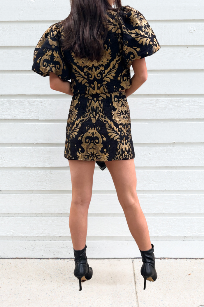 Black mini skirt with gold damask print and high waist, styled with puff sleeves and black ankle boots.