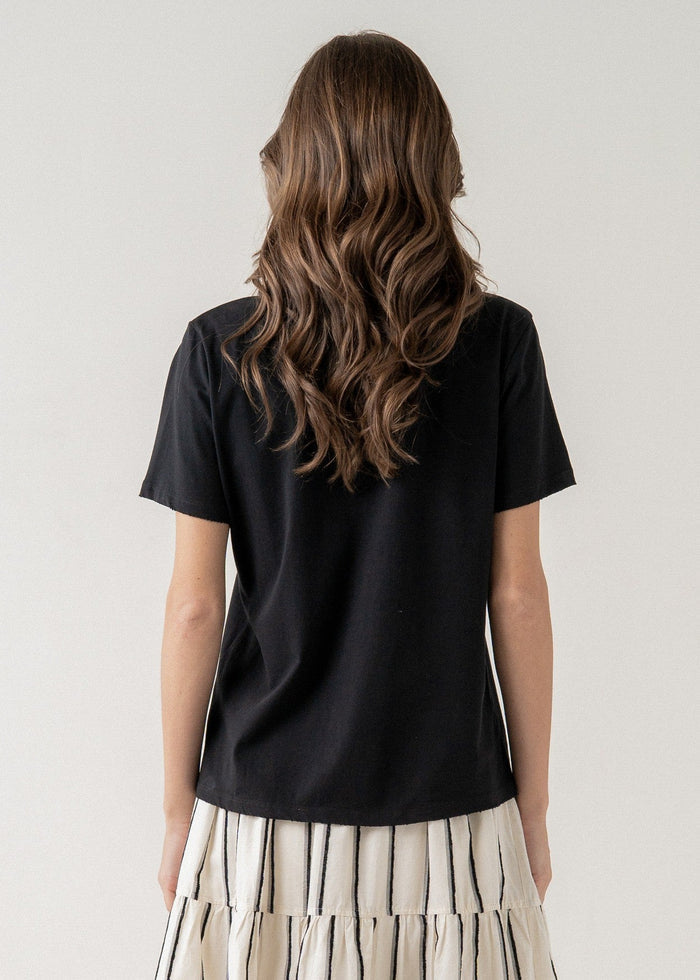 Black short-sleeve t-shirt with crew neckline, shown from back on model wearing cream pleated skirt.