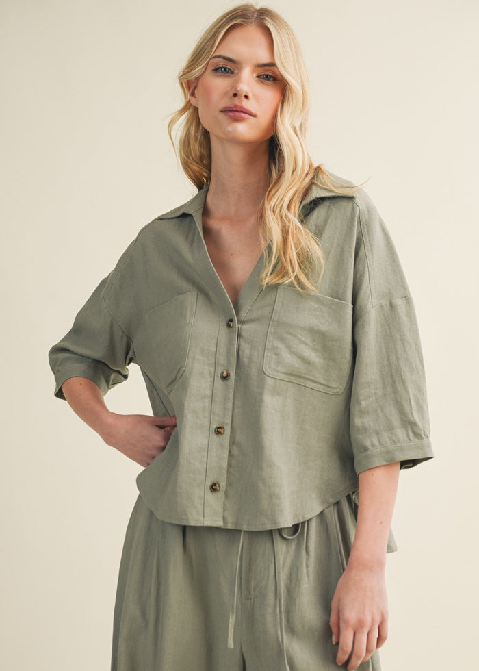 Sage green linen button-up top with three-quarter sleeves, chest pocket, and brown buttons on model.