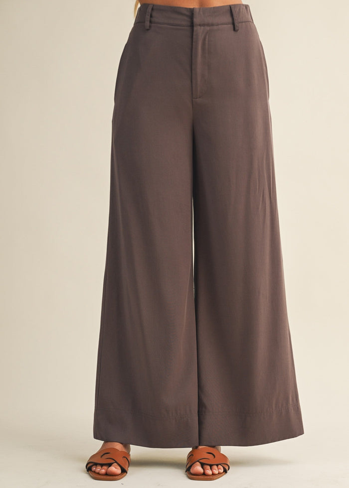 Brown wide-leg pants with zip fly and brown leather sandals on neutral background.