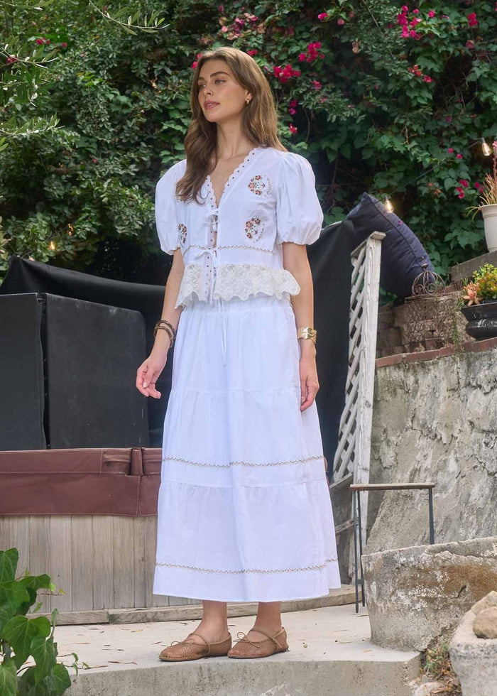 White midi skirt with tiered panels, lace trim details, and floral embroidery at waist.