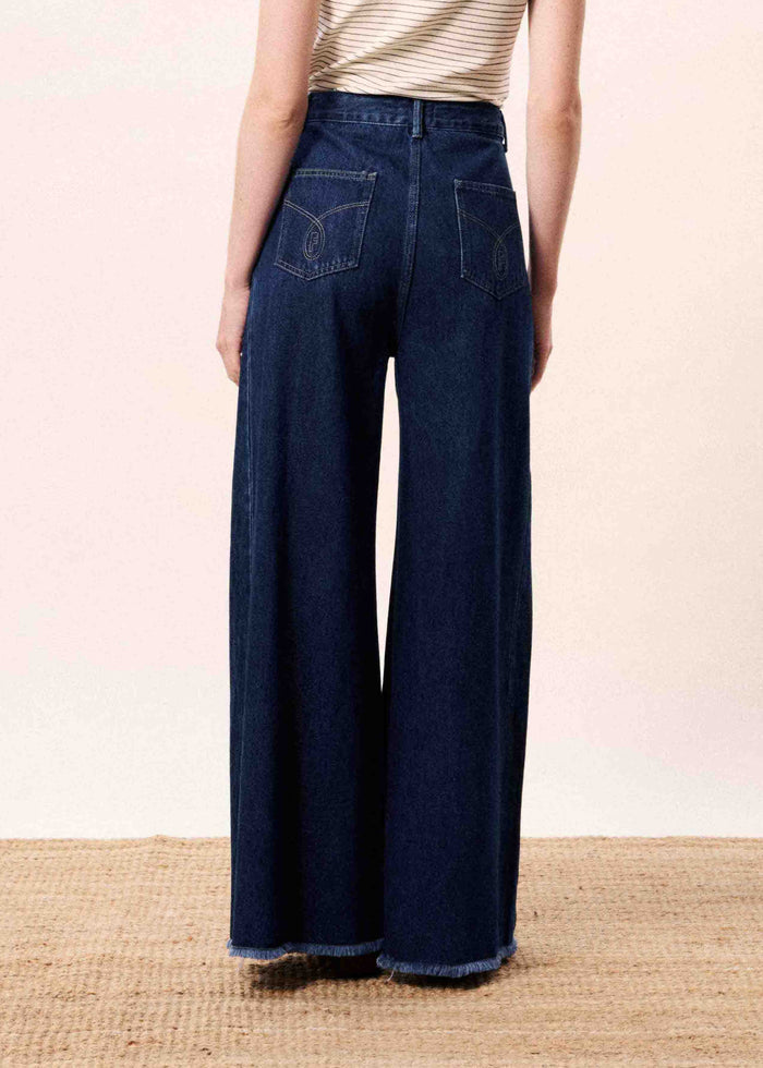 Woman wearing blue jeans standing in front of a white wall with a blue vase on a wooden surface.