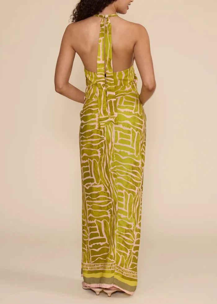 A woman shown from the back wearing a sleeveless halter-neck maxi dress with an open back and a bold abstract print in olive green and cream, set against a plain beige studio background.