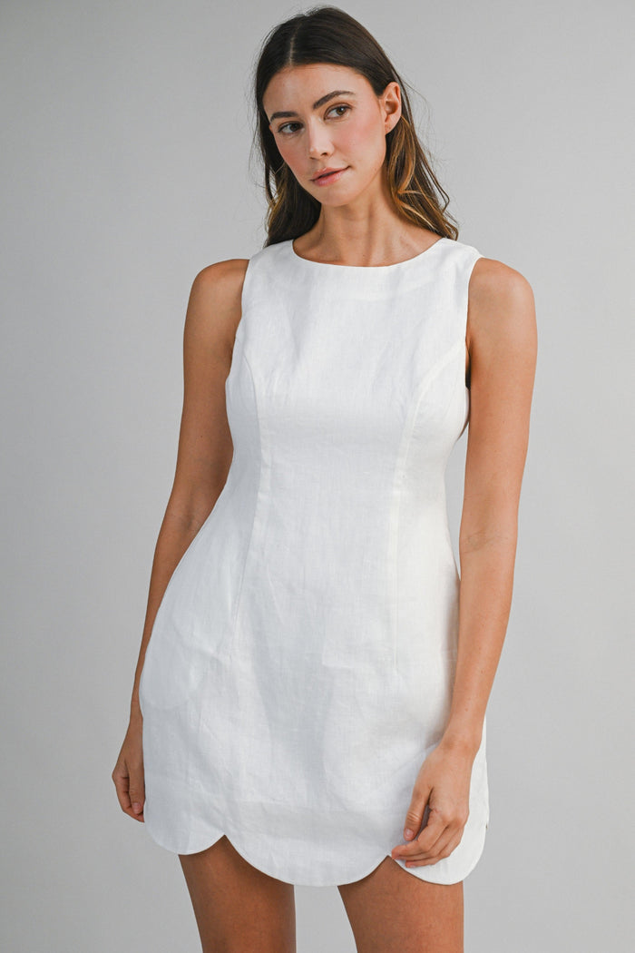 White sleeveless linen mini dress with rounded neckline and curved hemline on model.