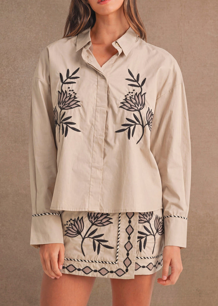 A woman modeling a beige long-sleeve button-up shirt with black floral embroidery and a matching embroidered mini skirt, set against a textured beige studio backdrop.