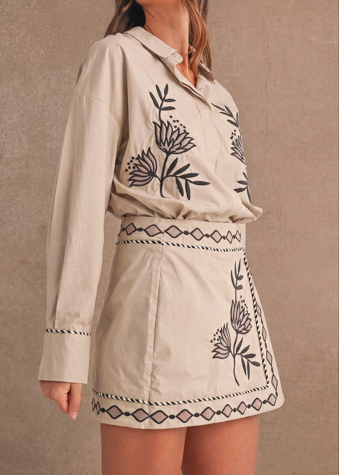 A woman modeling a beige long-sleeve shirt and matching mini skirt set with black embroidered botanical and geometric patterns, set against a textured beige wall.